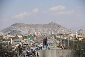 A wide cityscape of Kabul, Afghanistan, showing a busy highway with traffic, surrounded by buildings and a large mountain in the background under a cloudy sky.