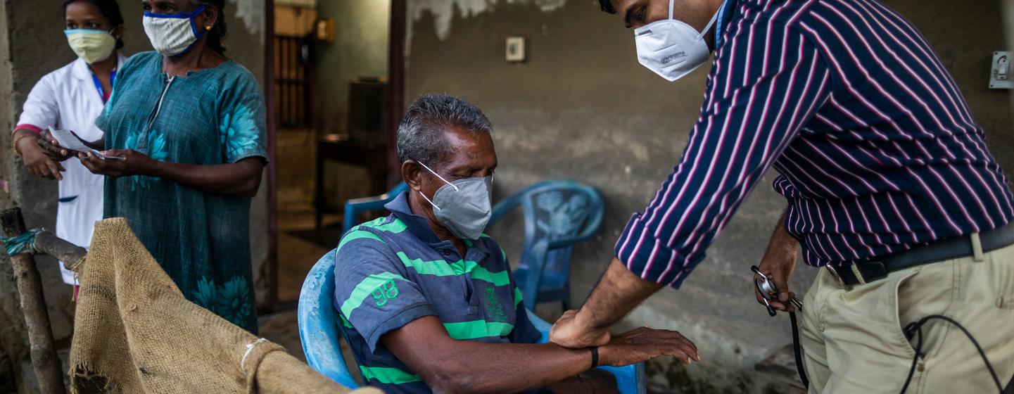 A masked doctor in a striped shirt examines the arm of a seated, masked elderly man during a vaccination session in a rustic room on Munroe Island, Kerala, while two women stand behind.