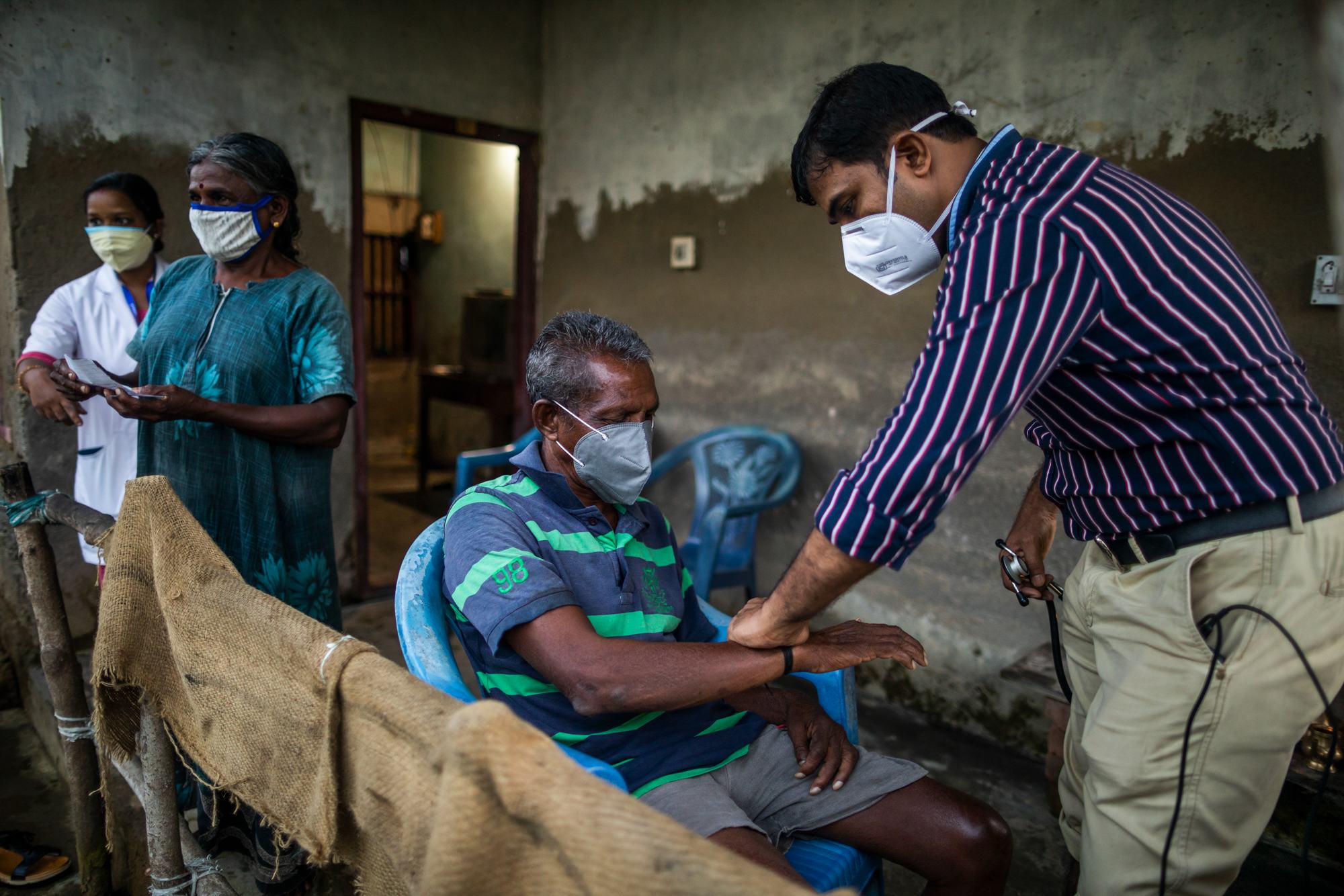 Un médico enmascarado con una camisa a rayas examina el brazo de un anciano sentado y también enmascarado durante una sesión de vacunación en una habitación rústica en la isla de Munroe, Kerala, mientras dos mujeres permanecen de pie detrás.