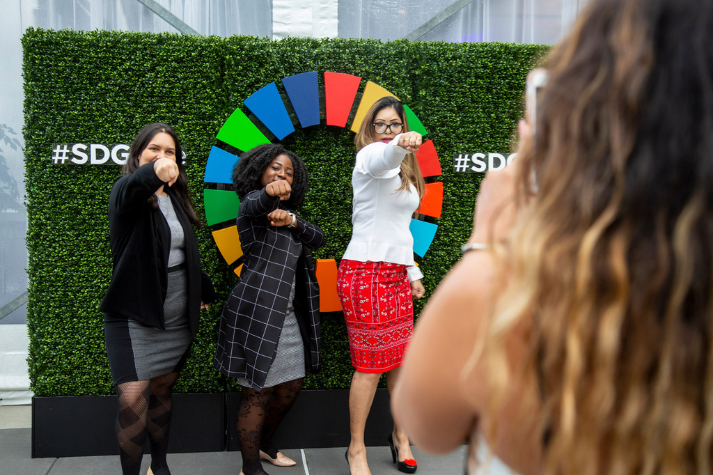 ثلاث شابات يتخذن وضعية لالتقاط صورة في فعالية تتميز بجدار أخضر يحمل وسم #SDG وعجلة SDG ملونة.