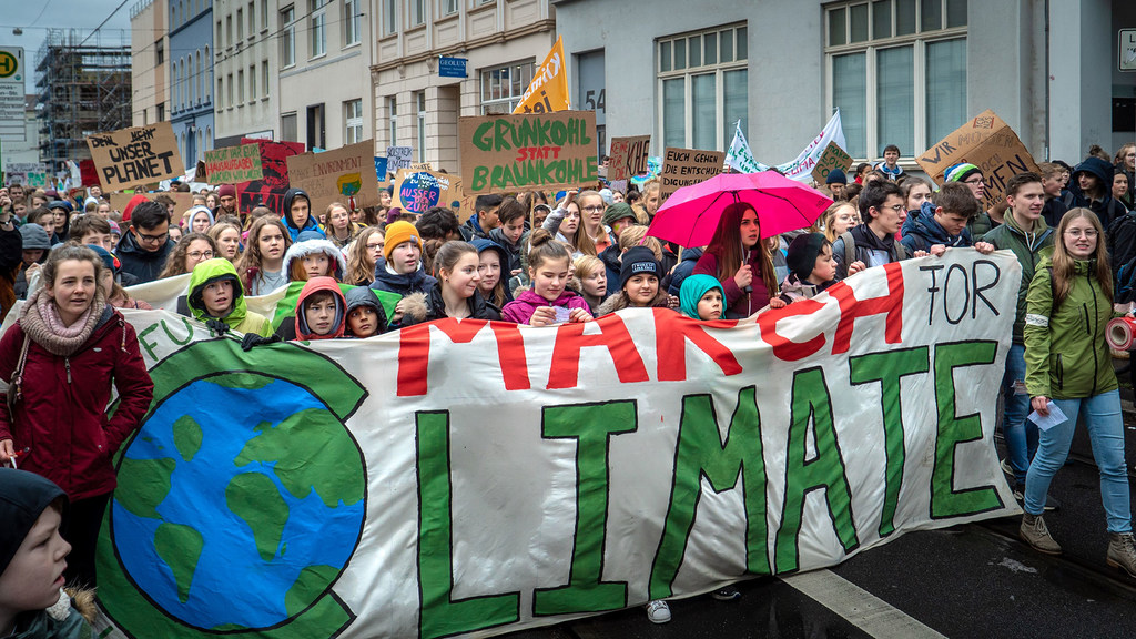 A large group of young climate activists marches in Bonn, Germany, carrying a banner that reads 'MARCH FOR CLIMATE'.