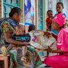 A health worker in pink UNFPA uniform presents medical supplies from a woven basket to a mother holding her baby, in a clinic setting in DR Congo.