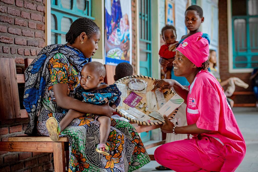 A health worker in pink UNFPA uniform presents medical supplies from a woven basket to a mother holding her baby, in a clinic setting in DR Congo.