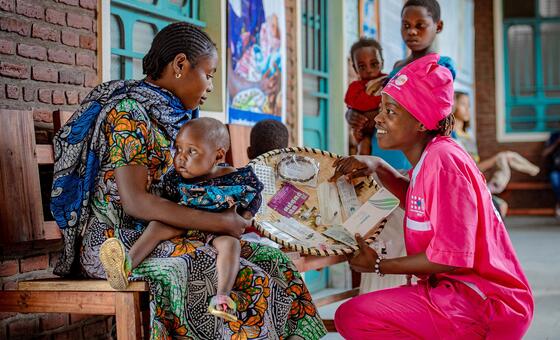 A health worker in pink UNFPA uniform presents medical supplies from a woven basket to a mother holding her baby, in a clinic setting in DR Congo.