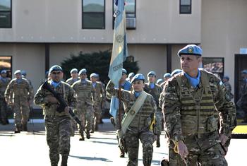 Soldados argentinos con uniformes de camuflaje y boinas azules marchan en fila, portando una bandera argentina y una bandera de la ONU durante una ceremonia de despedida.