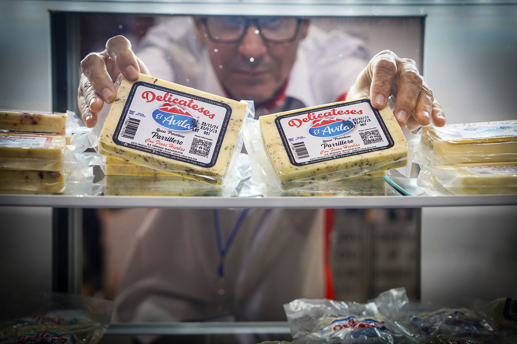 Un hombre con gafas y camisa blanca manipula paquetes de queso Parrillero de la marca El Ávila en una vitrina refrigerada.