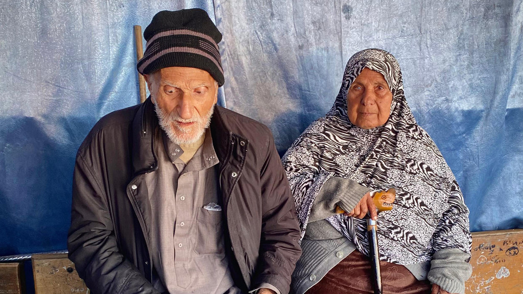 Mwanamume na mwanamke mzee wa Kipalestina, waliohamishwa kutoka Gaza, wamekaa ndani ya makazi ya UNRWA. Mwanamume amevaa koti na koti, huku mwanamke akiwa amevaa kitambaa cha kichwani chenye muundo na fimbo.
