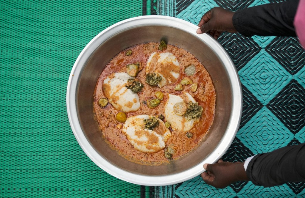 Mtu anashikilia bakuli kubwa la chuma lenye kitoweo chenye vipande vya kuku na uji mgumu wa Fonio, uliowekwa kwenye mkeka wenye muundo.