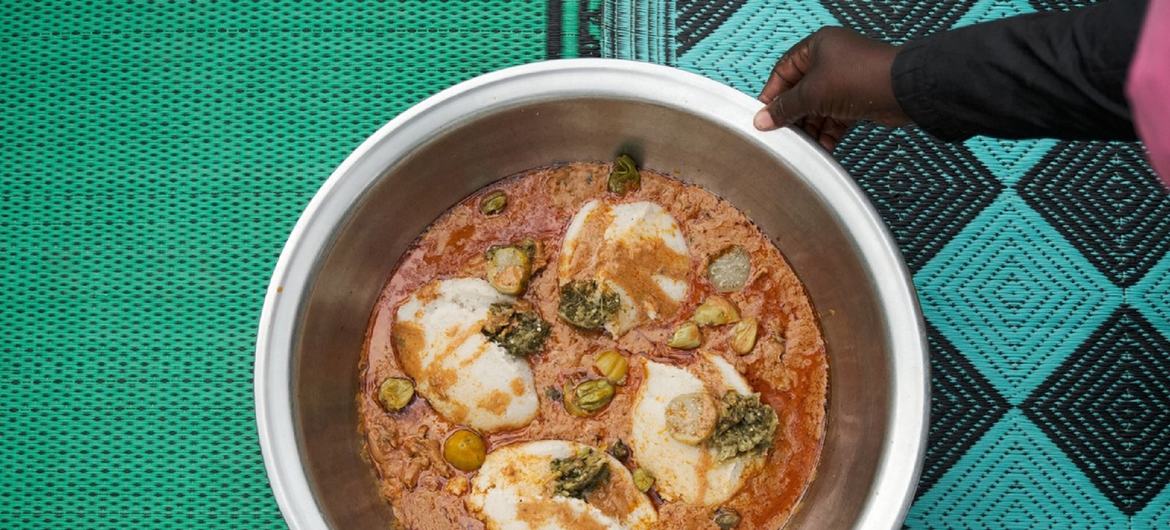 Человек держит большую металлическую миску с тушеным мясом, кусочками курицы и густой кашей из фонио, поставленную на узорчатую циновку.