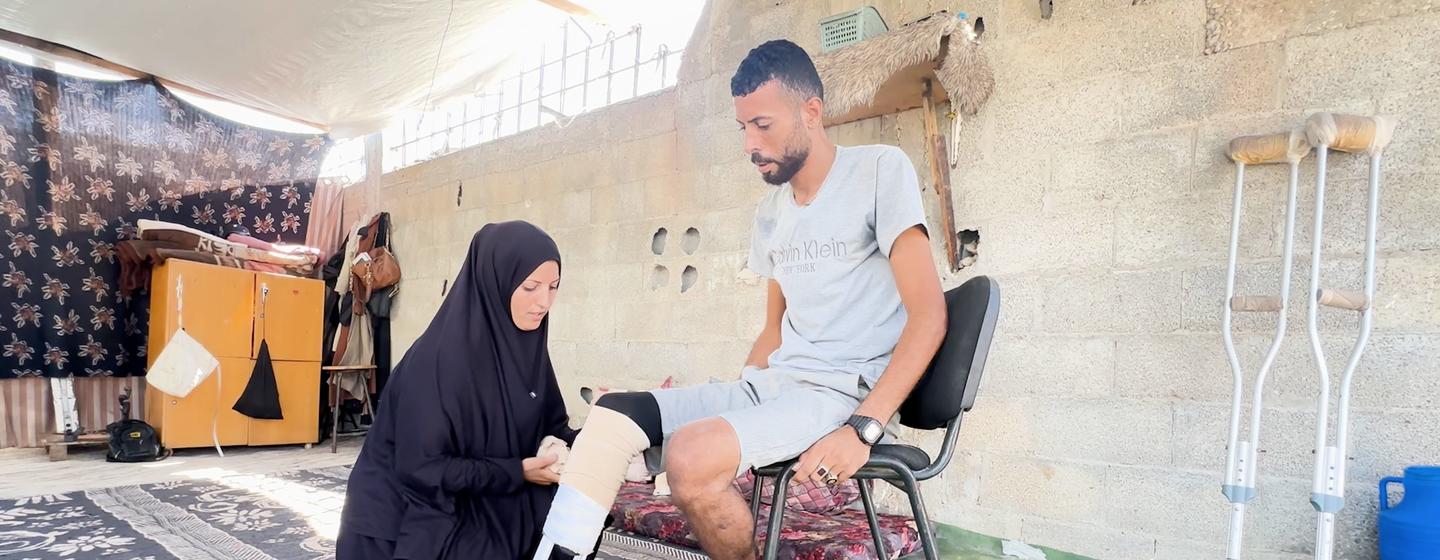 Ibrahim Abdel Nabi, un palestino, está sentado en una silla mientras su esposa le ayuda a ponerse una prótesis de pierna hecha a mano. Al fondo se ven unas muletas.
