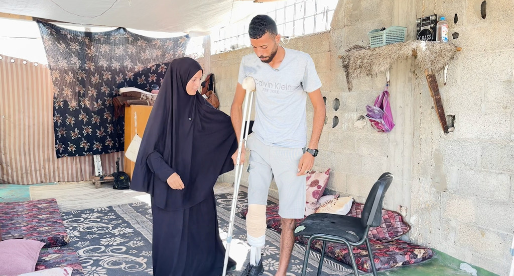 إبراهيم عبد النبي، فلسطيني نازح في غزة، يمشي باستخدام طرفه الاصطناعي المصنوع يدوياً، بينما تساعده زوجته.