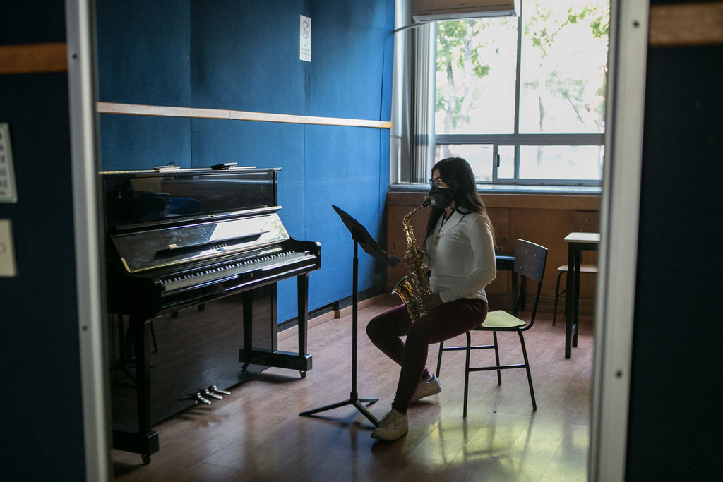 Una mujer con gafas y mascarilla está sentada en una silla en una sala de música, tocando el saxofón junto a un piano.