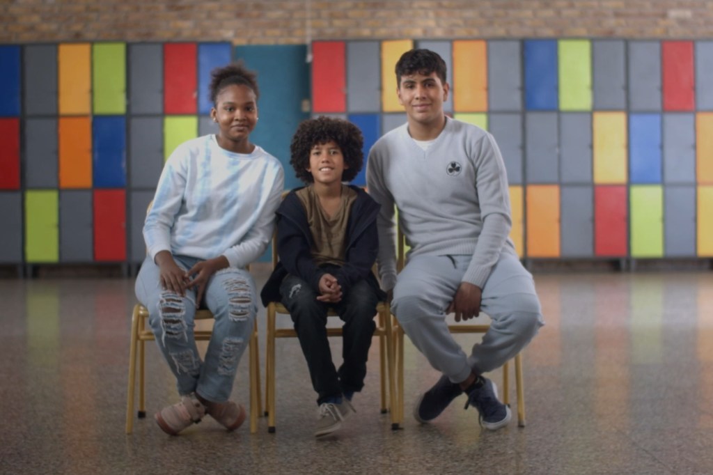Três estudantes afrodescendentes de diferentes idades sentam-se em cadeiras no corredor de uma escola, com uma parede colorida de armários ao fundo.