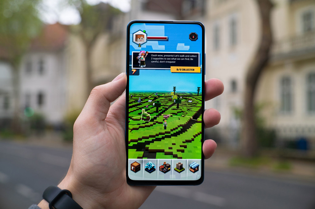Uma mão segura um smartphone exibindo o jogo de realidade aumentada Minecraft Earth em uma rua urbana.