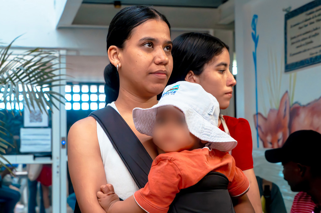 Una mujer venezolana sostiene a un niño pequeño en un portabebés mientras espera en el interior de una casa.