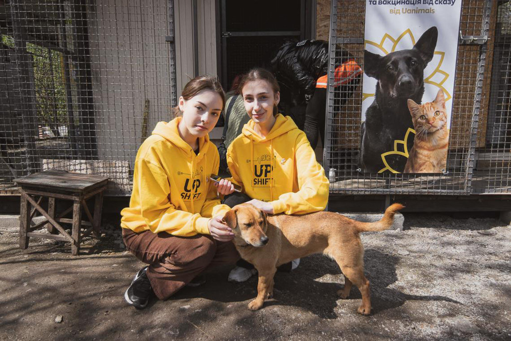 Two young women in yellow hoodies kneel and pet a light brown dog in front of an animal shelter in Ukraine.