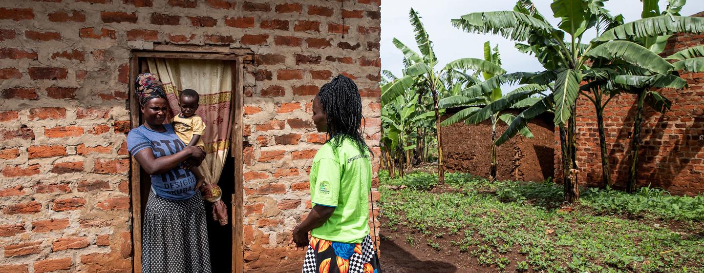 Une femme vêtue d'une chemise bleue et d'une jupe à motifs, tenant un bébé dans les bras, se tient sur le seuil d'une maison en briques tandis qu'une autre femme, portant une chemise verte et une jupe colorée, s'approche d'elle.