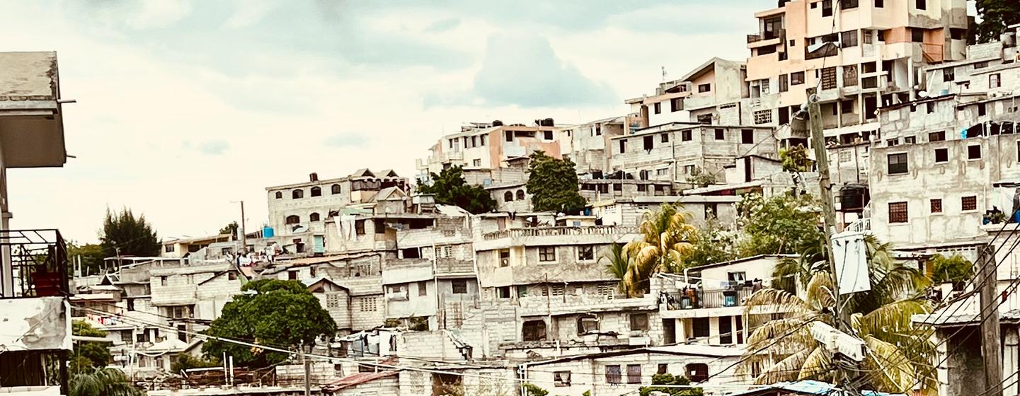 A street scene in the Delmas commune of Port au Prince, Haiti, with a black Nissan Pathfinder and a motorcycle in the foreground, and densely packed houses built on a hillside in the background.