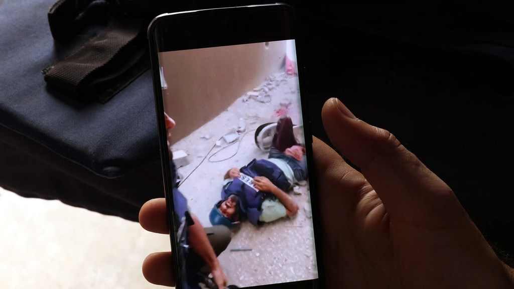 Primer plano de una mano que sostiene un teléfono inteligente que muestra un video de un hombre herido en el suelo, en una zona llena de escombros.