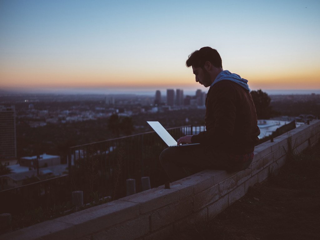 Un hombre está sentado en un muro usando una computadora portátil al atardecer, con el perfil urbano de la ciudad de fondo.