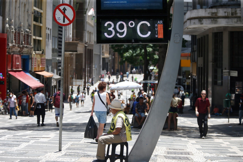 Um homem de colete amarelo e chapéu senta-se num banquinho numa calçada quente e ensolarada de São Paulo, no Brasil, com um grande sinal de termômetro indicando 39°C.