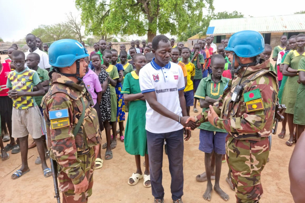 Walinda amani wawili UNMISS wa Bangladesh waliovalia kofia za bluu wakipeana mikono na mwanamume wa eneo hilo mbele ya kundi la watoto huko Tonj, Sudan Kusini.