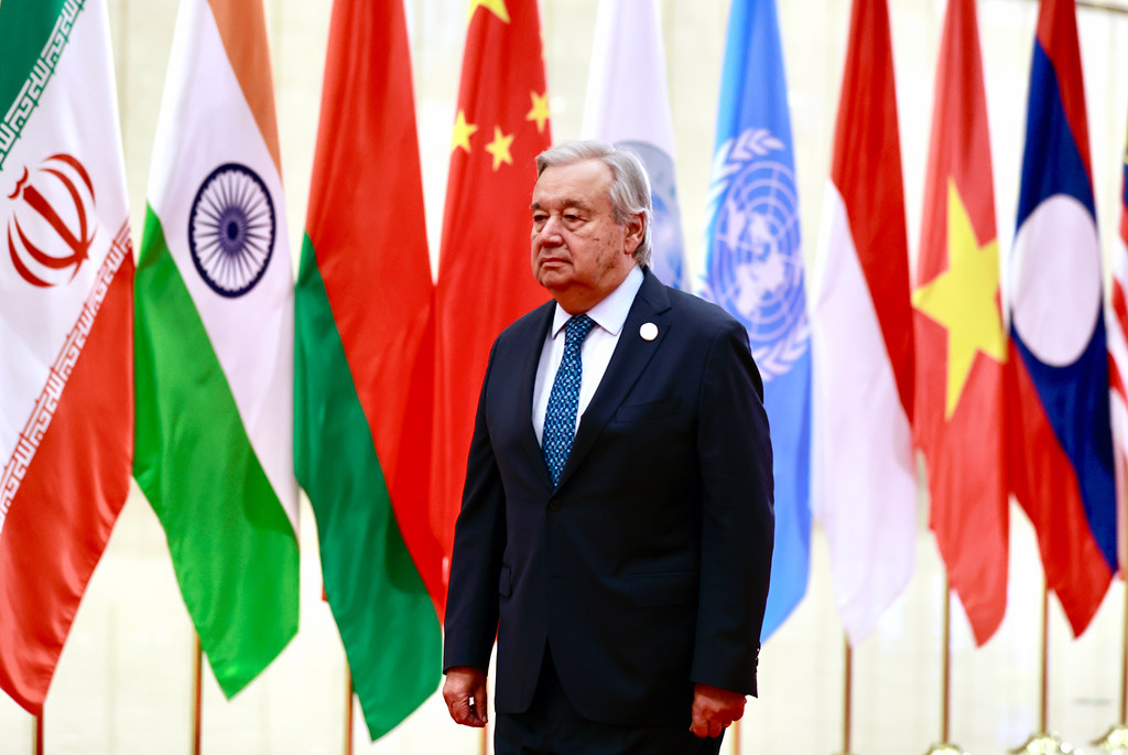 Katibu Mkuu UN António Guterres anatembea mbele ya safu ya bendera za kimataifa, ikiwa ni pamoja na zile za Iran, India, China, na UMOJA WA MATAIFA.
