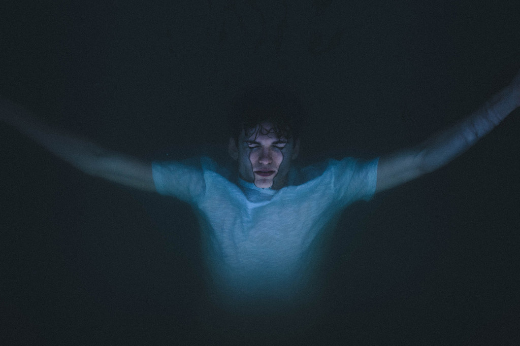 Um homem com os braços estendidos está submerso em água escura, iluminado por uma luz azul vinda de baixo.