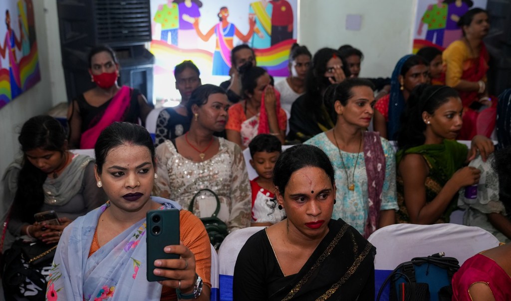 एक इनडोर सभा में ट्रांसजेंडर महिलाओं का एक समूह और एक बच्चा बैठे हैं, जिनमें से एक महिला के हाथ में स्मार्टफोन है।
