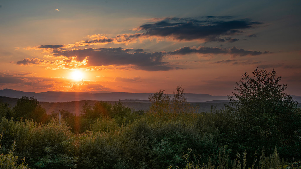 Una puesta de sol sobre un paisaje montañoso en Rumania, con el sol ocultándose tras las nubes y las montañas.