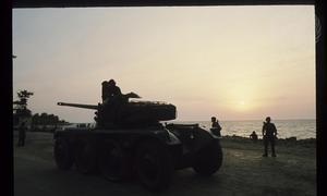 Um veículo blindado militar e soldados são silhuetados contra um pôr do sol sobre o oceano em Angola.