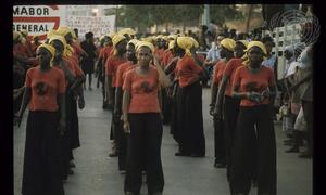 Um grupo de mulheres angolanas vestindo camisetas vermelhas e lenços amarelos na cabeça marcha em formação por uma rua.