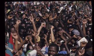 Uma multidão de pessoas com os punhos cerrados celebra um evento em Angola, com o emblema da ONU visível ao fundo.