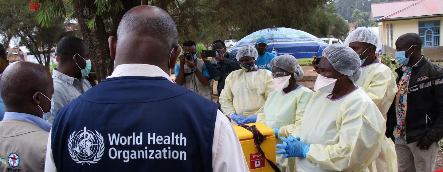 Un groupe de membres du personnel médical de l'Organisation mondiale de la santé, vêtus de tenues de protection, se tient à côté d'une boîte jaune de signalisation biologique lors d'une intervention contre le virus Ebola en Guinée ou en République démocratique du Congo.