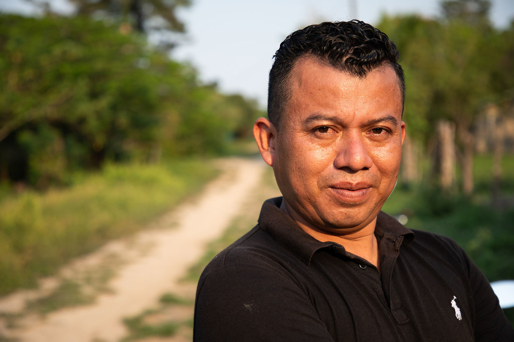 Retrato de un hombre con una camisa polo negra de pie en un camino de tierra en un entorno rural.