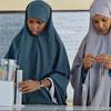 Wanafunzi wawili wa kike waliovaa hijabu wanafanya jaribio la sayansi darasani. Mwanafunzi aliye upande wa kushoto anamimina kioevu kutoka kwenye kopo kwenye silinda iliyokamilika, huku mwanafunzi aliye upande wa kulia akishika kamba.
