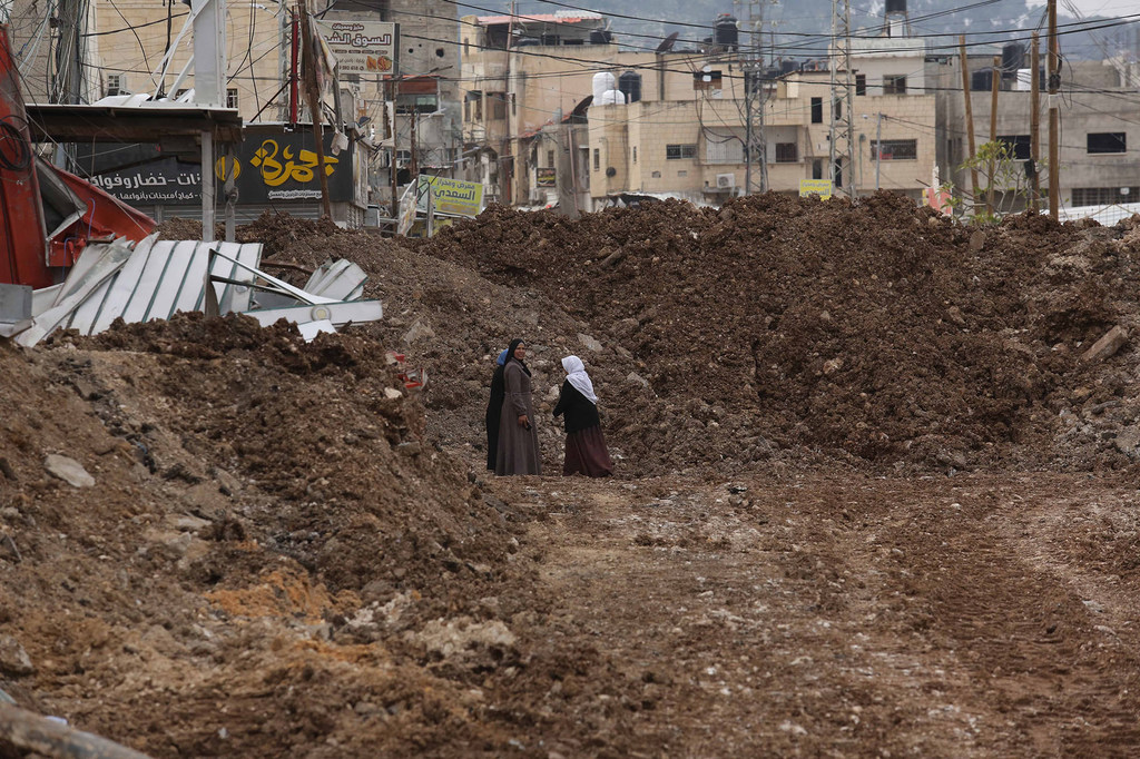 Mujeres palestinas permanecen de pie en un camino embarrado y excavado en Jenin, Cisjordania, con montones de escombros de edificios destruidos al fondo.