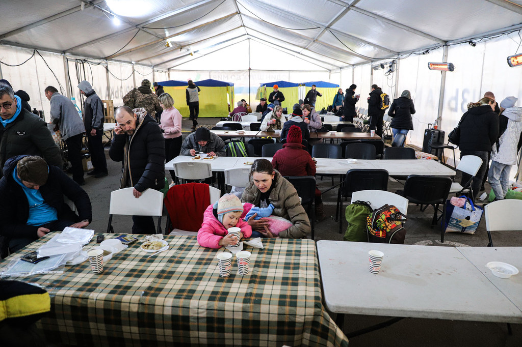 En abril de 2022, personas desplazadas internamente, entre ellas mujeres y niños, están sentadas a mesas dentro de un centro de acogida en Zaporiyia, Ucrania.