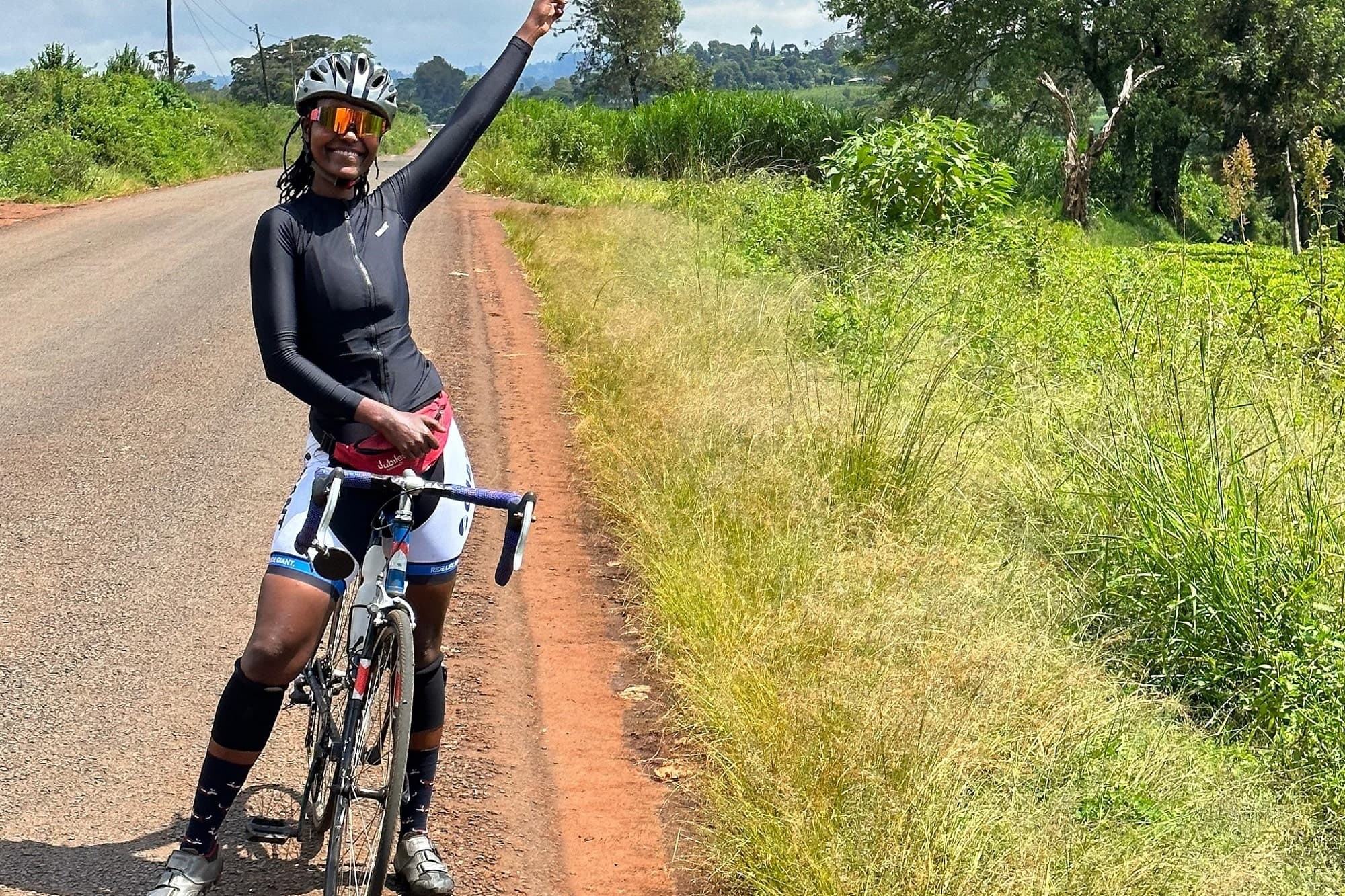 Muthoni, mwanamke anayeendesha baiskeli, amesimama na baiskeli yake kwenye barabara ya vijijini nchini Kenya, akiinua mkono wake kama ishara ya salamu au ushindi.