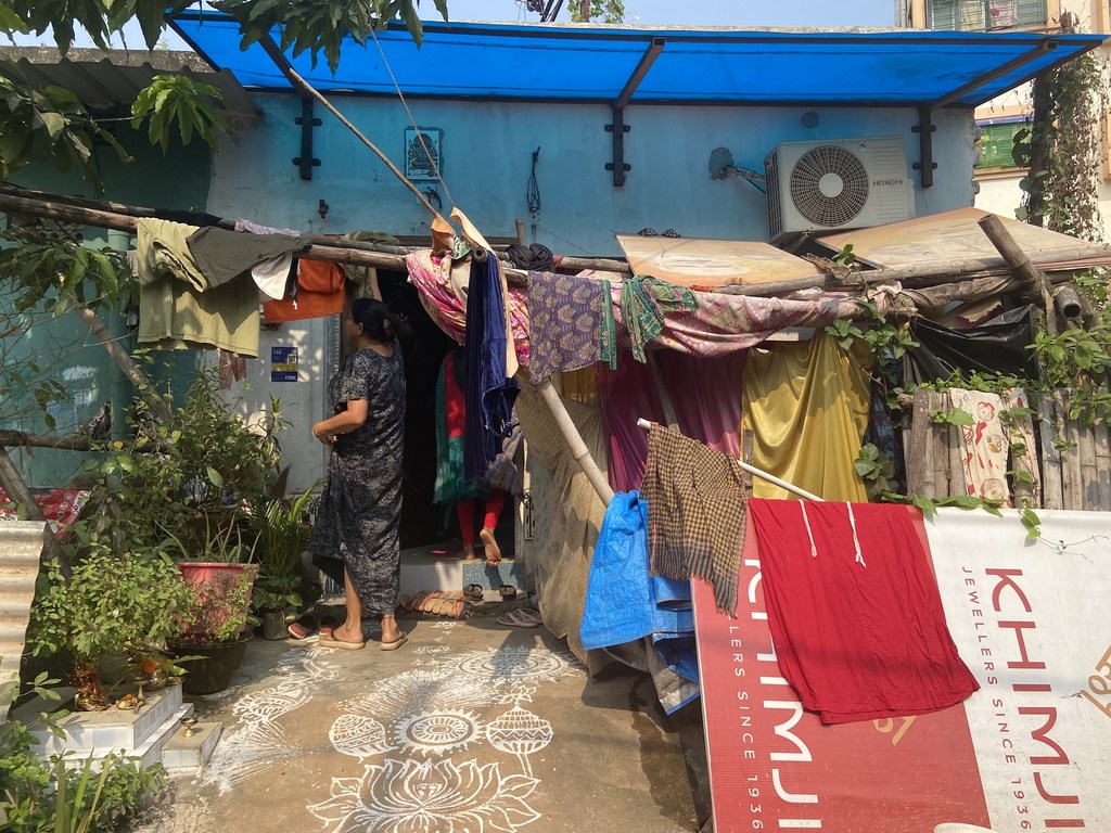 एक महिला भारत की एक अनौपचारिक बस्ती में नीली तिरपाल की छत वाले एक अस्थायी घर के दरवाजे पर खड़ी है।