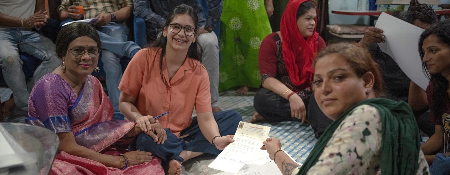 एक सामुदायिक बैठक में, ट्रांसजेंडर व्यक्तियों सहित महिलाओं का एक समूह फर्श पर बैठकर आवास के वास्तुशिल्पीय योजनाओं की जांच कर रहा है।