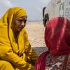 Mfanyakazi wa kibinadamu mwanamke aliyevaa hijabu ya njano akizungumza na mhamiaji mwanamke aliyevaa hijabu nyekundu karibu na gari la IOM jangwani.