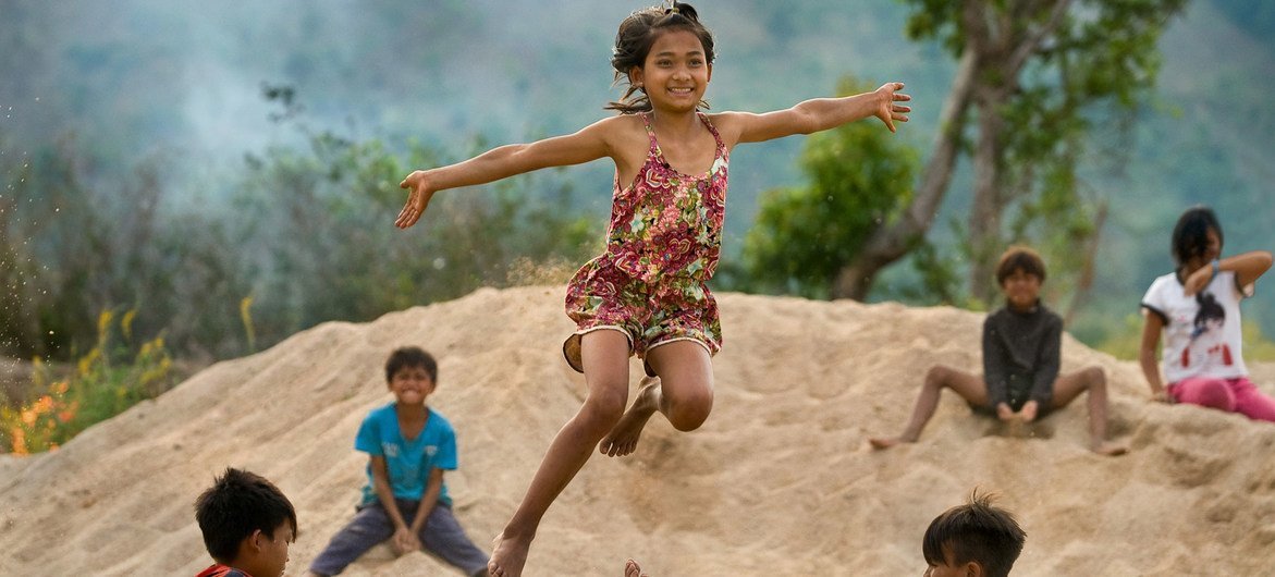 Una niña alegre salta por el aire sobre dos niños sentados en un montículo de arena, rodeados de otros niños en un paisaje rural vietnamita.