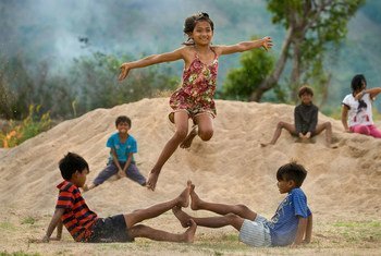 Una niña alegre salta por el aire sobre dos niños sentados en un montículo de arena, rodeados de otros niños en un paisaje rural vietnamita.