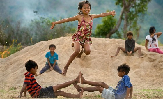 Una niña alegre salta por el aire sobre dos niños sentados en un montículo de arena, rodeados de otros niños en un paisaje rural vietnamita.