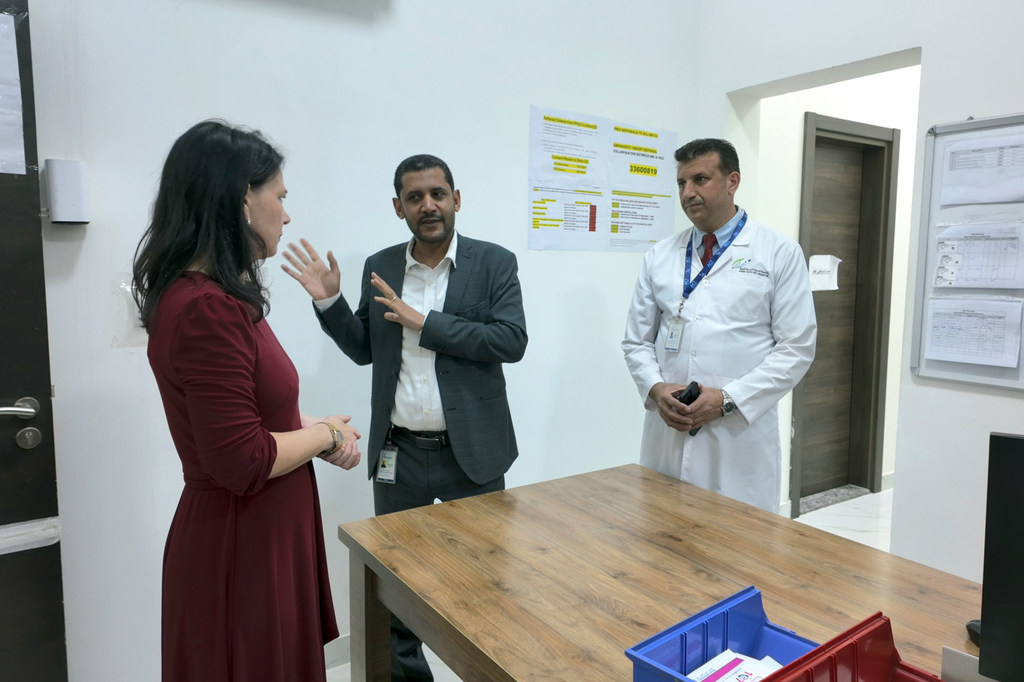 امرأة ترتدي فستاناً عنابي اللون تتحدث مع رجلين، أحدهما يرتدي بدلة والآخر معطفاً أبيض، داخل عيادة طبية.