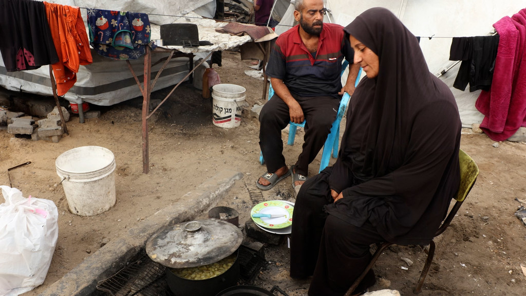 امرأة ورجل يجلسان خارج مأوى مؤقت في مخيم للاجئين، مع قدر من الطعام يطهى على نار مكشوفة.