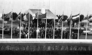 Uma multidão se reúne em frente a um prédio com diversas bandeiras hasteadas, incluindo a de Angola. Repórteres e fotógrafos estão presentes no evento.