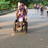 Um homem em cadeira de rodas participa de uma maratona, empurrado por um acompanhante. Outros corredores são visíveis na estrada durante o evento.