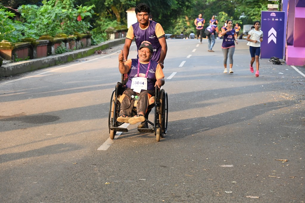 एक व्यक्ति व्हीलचेयर पर बैठकर मैराथन में भाग ले रहा है, जिसे उसका साथी धक्का दे रहा है। दौड़ के दौरान सड़क पर अन्य धावक भी दिखाई दे रहे हैं।